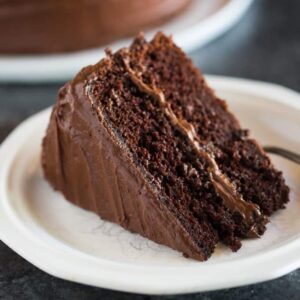 A slice of Hershey's perfectly chocolate chocolate cake, with chocolate frosting, on a white plate with a chocolate cake in the background.