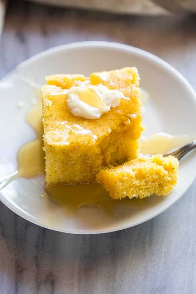 A piece of cornbread on a plate with a piece of butter on top, syrup on top and a bite taken out by a fork resting on the plate.