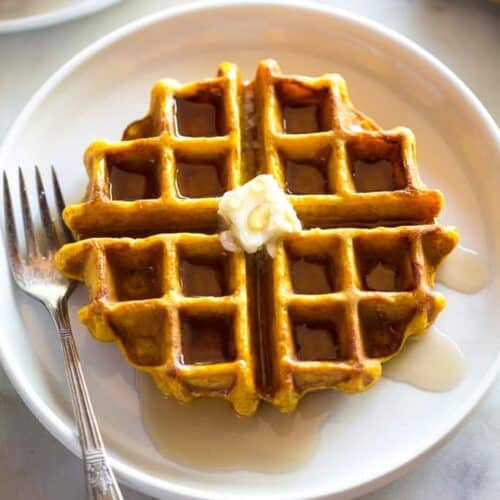 A pumpkin waffle with a syrup and a slice of butter on top, on a white plate with a fork, fall leaves and another plate of waffles to the side.