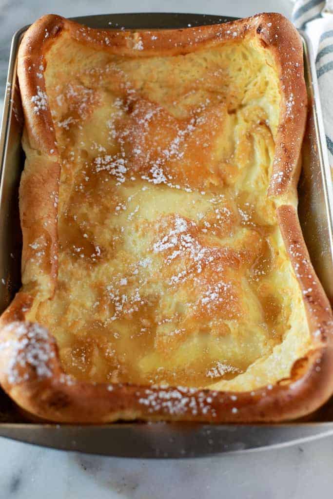 A metal 9x13 inch pan with baked german pancakes in it, sprinkled with powdered sugar and syrup.