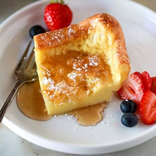 A slice of German Pancakes on a white plate with powdered sugar and syrup on top, served with strawberries and blueberries.