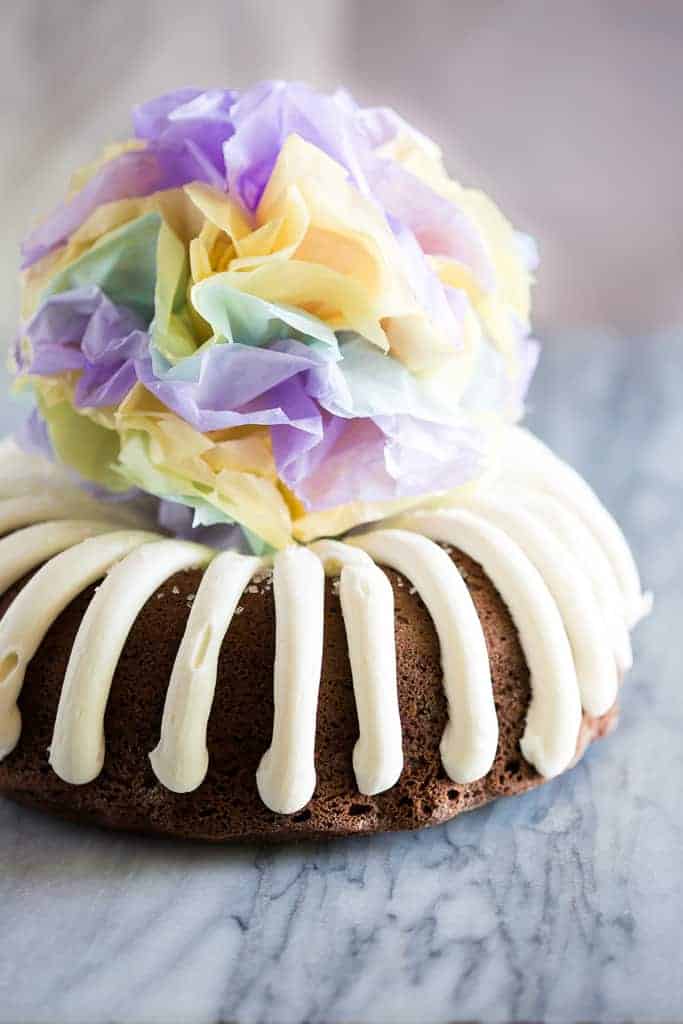 A chocolate bundt cake with cream cheese frosting and a tissue paper pom pom in the center of the cake.