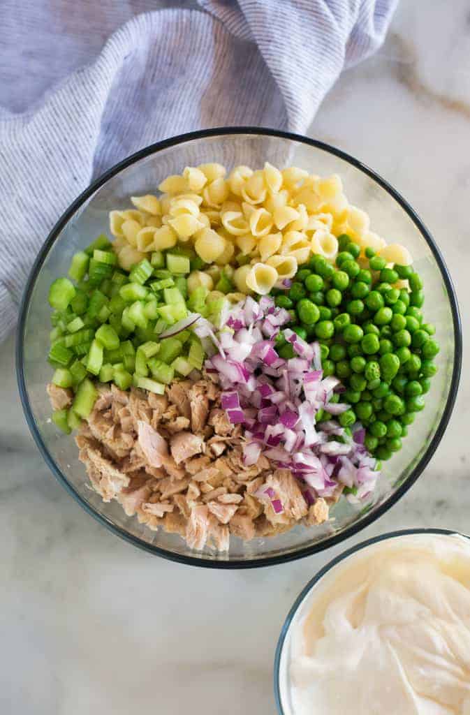 A clear glass mixing bowl with the ingredients for tuna pasta salad including cooked pasta, celery, onion, peas and tuna, next to a small bowl with a greek yogurt and mayonnaise sauce.