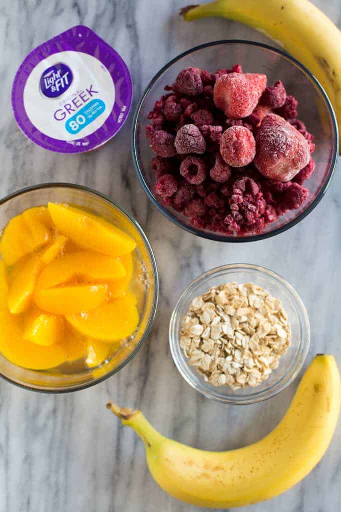 The ingredients needed to make a healthy breakfast smoothie, including frozen berries, Greek yogurt, canned peaches, bananas and rolled oats.