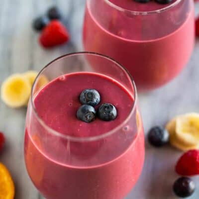 Two clear glass cups filled with a healthy breakfast smoothie with three blueberries placed on top and extra fruit in the background.