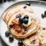 Three blueberry pancakes on a plate, with a slab of butter on top, and fresh blueberries.