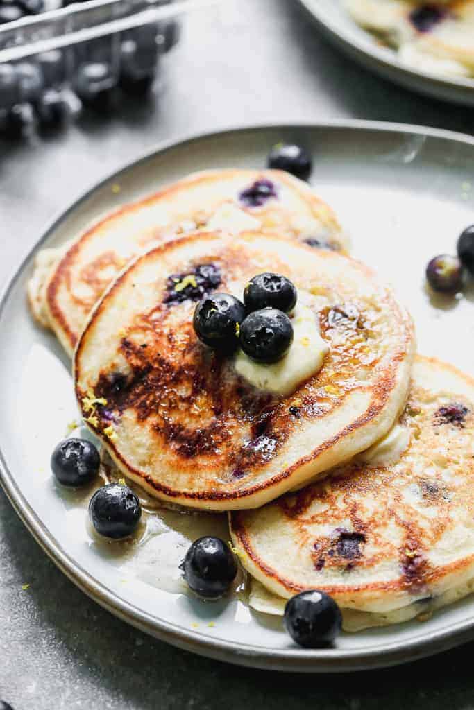 Three blueberry pancakes on a plate, with a slab of butter on top, and fresh blueberries.