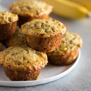 Banana Bran Muffins stacked on a white plate with bananas in the background.