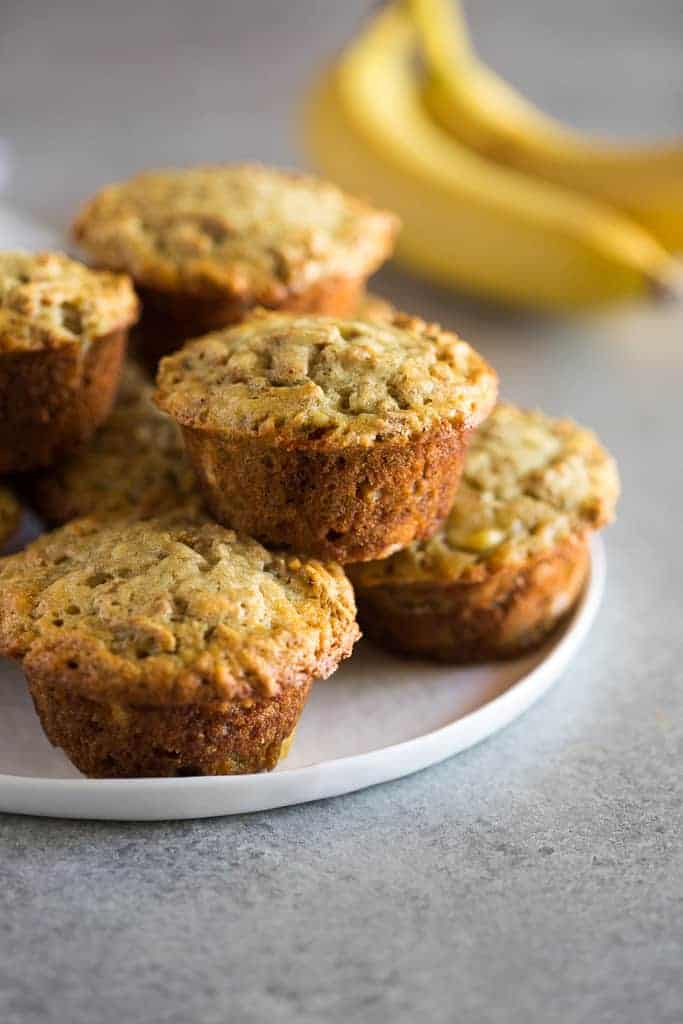 Banana Bran Muffins stacked on a white plate with bananas in the background.