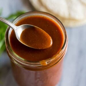 A mason jar filled with homemade enchilada sauce and a spoon lifting sauce up.