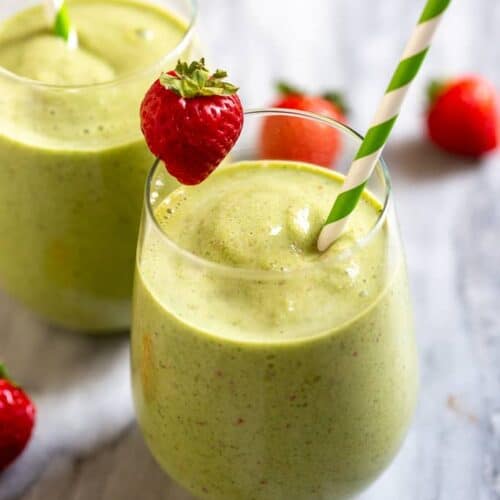 Green smoothie in two cups with a straw and strawberry on the edge of the cups.