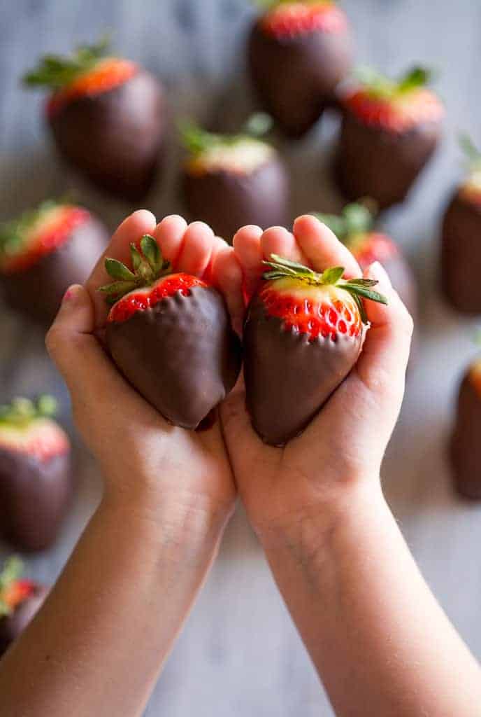 Two hands cupped together with two chocolate covered strawberries in them and chocolate covered strawberries in the background.