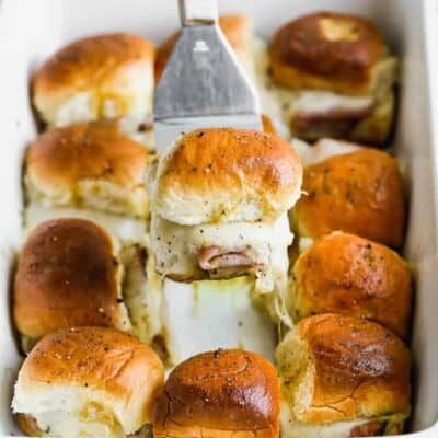 A spatula lifting a baked ham and cheese slider out of a white baking dish filled with sliders.