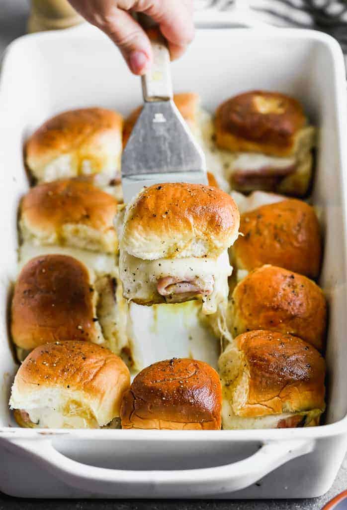 A spatula lifting a baked ham and cheese slider out of a white baking dish filled with sliders.