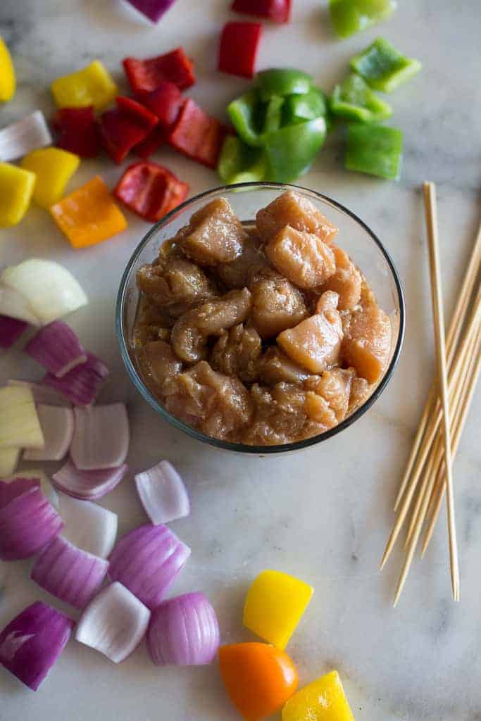 The ingredients for chicken kebabs included chunks of marinated chicken in a bowl surrounded by chopped purple onion, bell pepper and bamboo skewers.