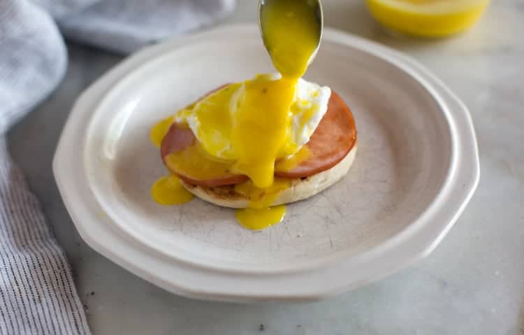 Homemade hollandaise sauce being spooned over a poached egg, canadian bacon and an English muffin to make eggs benedict.