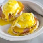 A plate with two eggs benedict which includes english muffin topped with a slice of canadian bacon, poached eggs and homemade hollandaise sauce.