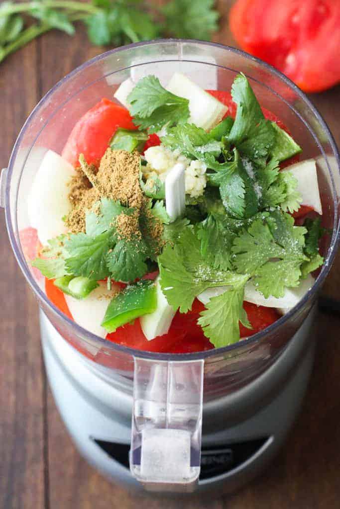 A food processor with the ingredients to make salsa in it, including tomatoes, cilantro, onion, garlic and cumin.