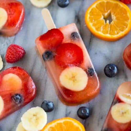 Homemade popsicles made with blueberries, strawberries, bananas and juice, laying on a marble board with fresh fruit scattered around them.