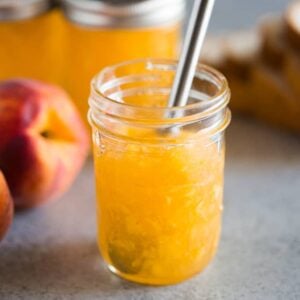 An open jar of peach jam with a butter knife in it and three full jars of jam and a whole peach in the background.