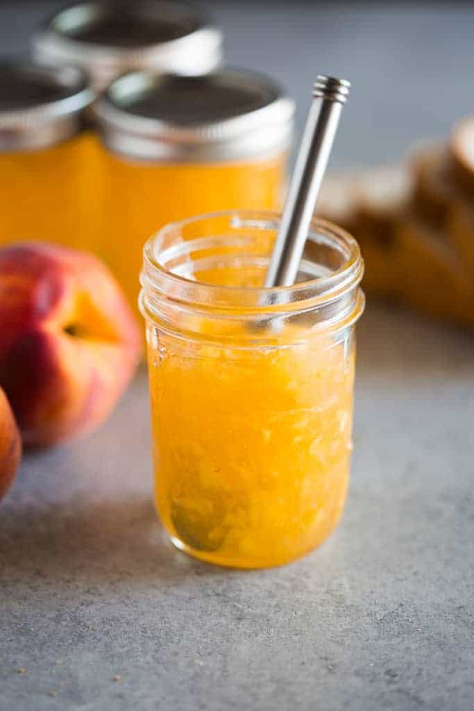 An open jar of peach jam with a butter knife in it and three full jars of jam and a whole peach in the background.