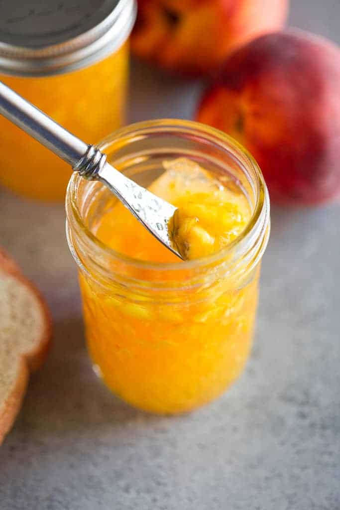 Overhead photo of a butter knife lifting fresh peach jam from a jar and a piece of bread, a peach and another jar of jam in the background.