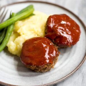 Two mini meatloaf muffins with meatloaf sauce on them on a plate with mashed potatoes and green beans.