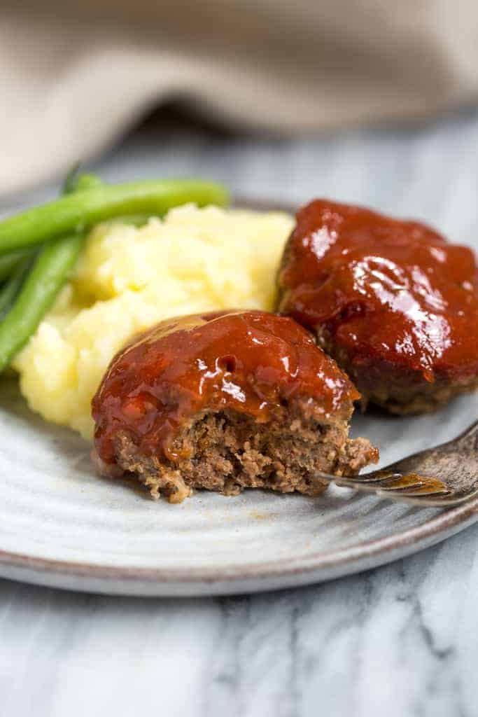 A mini meatloaf muffin on a plate with a bite taken out of it and another meatloaf, mashed potatoes and green beans in the background.