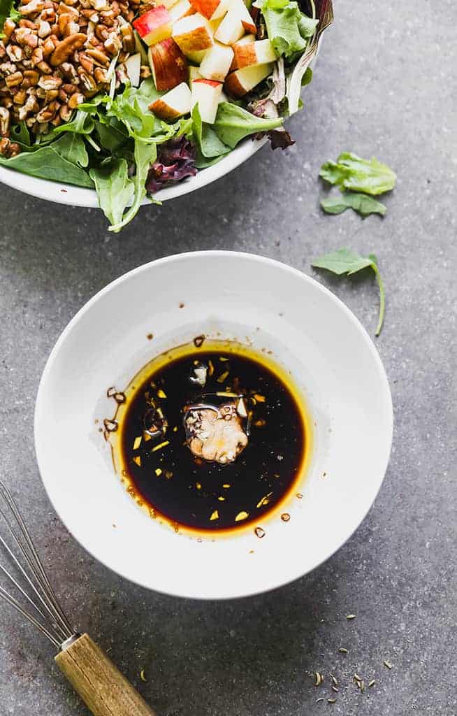 A white bowl with balsamic vinegar, mustard and oil in it to mix for dressing a salad in the background.