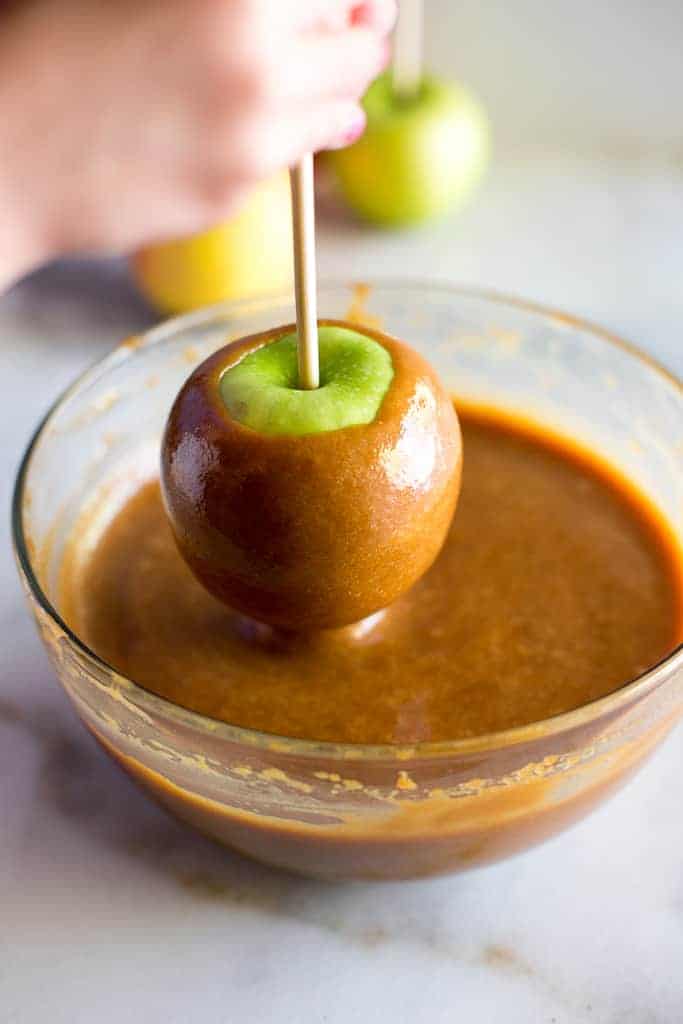 A hand holding a stick with a green apple on it and dipping it into a bowl full of melted caramel.