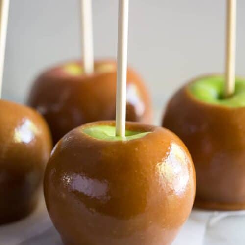 Four homemade caramel apples with a stick in them, on parchment paper on a white board.