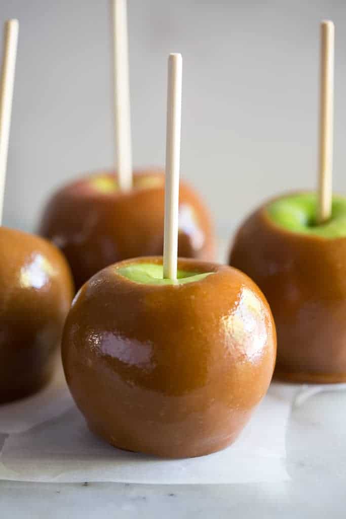 Four homemade caramel apples with a stick in them, on parchment paper on a white board.