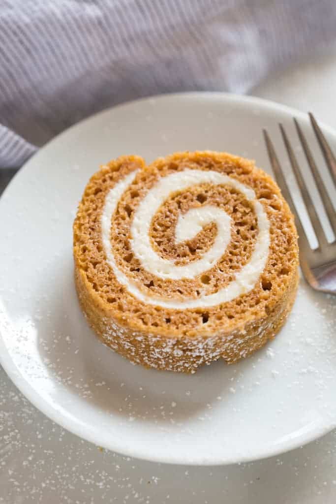 One round slice of a pumpkin roll on a white plate with sprinkled powdered sugar and a fork.