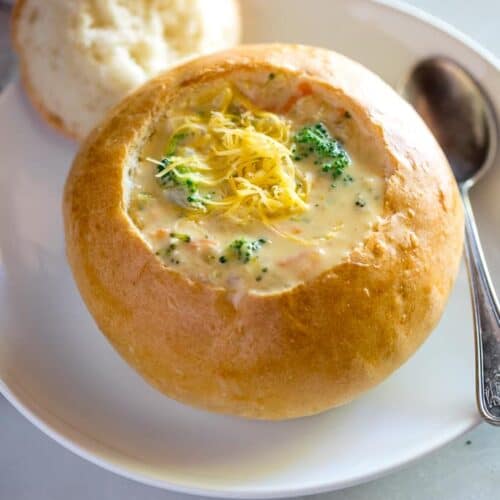A homemade bread bowl on a white plate with the center cut out, resting on the edge of the plate, broccoli cheese soup in the bread bowl and a spoon on the side.