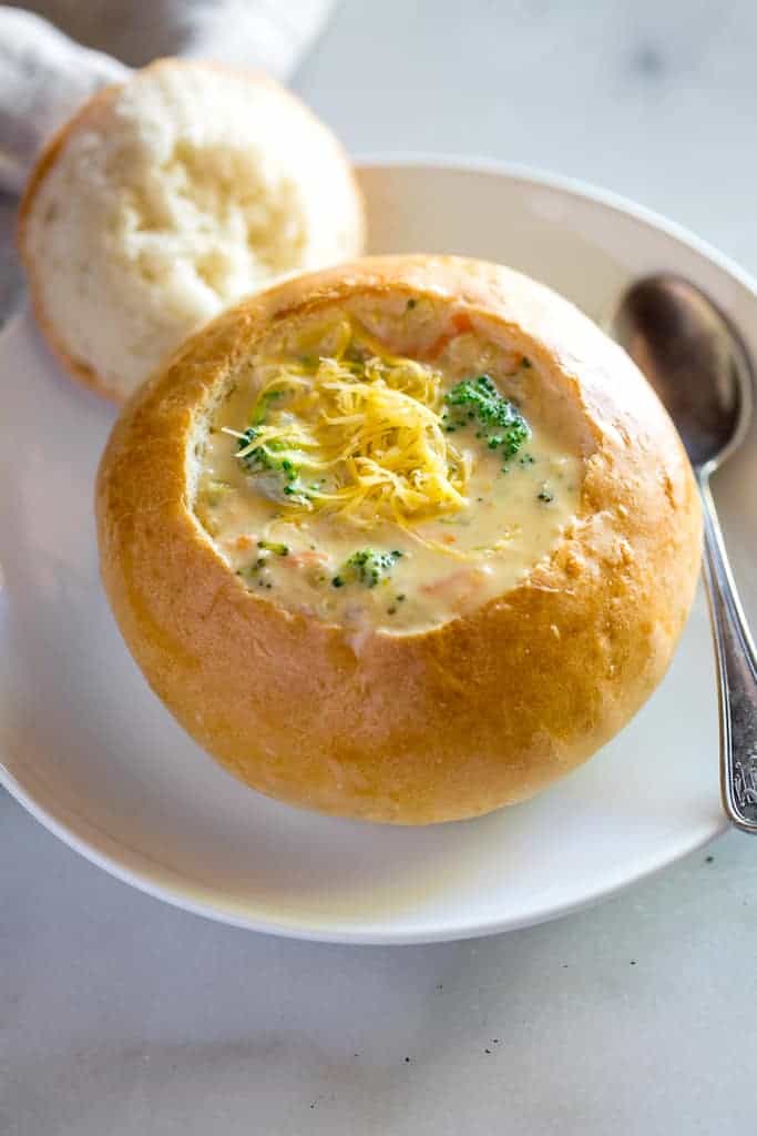 A homemade bread bowl on a white plate with the center cut out, resting on the edge of the plate, broccoli cheese soup in the bread bowl and a spoon on the side. 