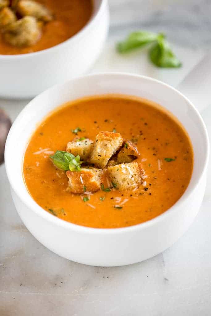 A white bowl full of tomato soup with croutons as a garnish and another bowl of soup in the background.