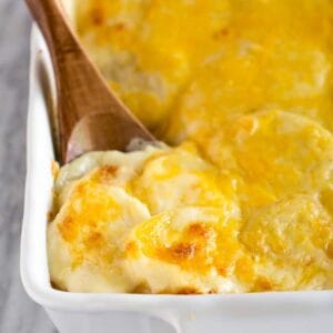 A white casserole dish with baked Potatoes Au Gratin and a wooden spoon to serve them.