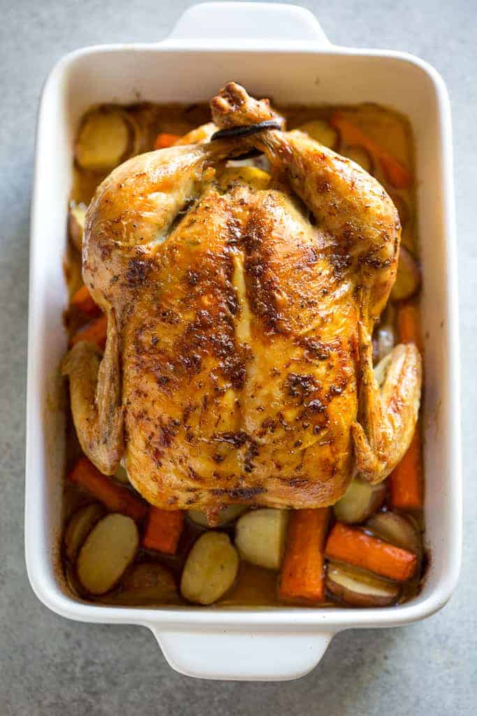 Overhead view of a roast chicken on top of chopped potatoes, carrots and onion, in a white baking dish.
