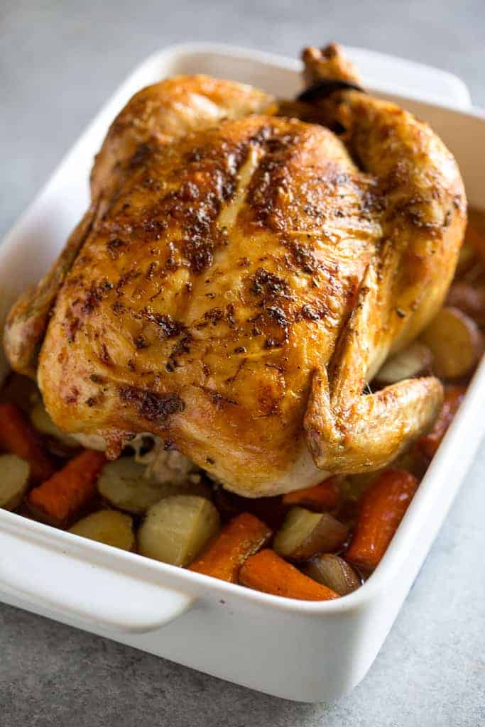 A roasted chicken on a bed of vegetables in a white 9 by 13 inch baking dish.