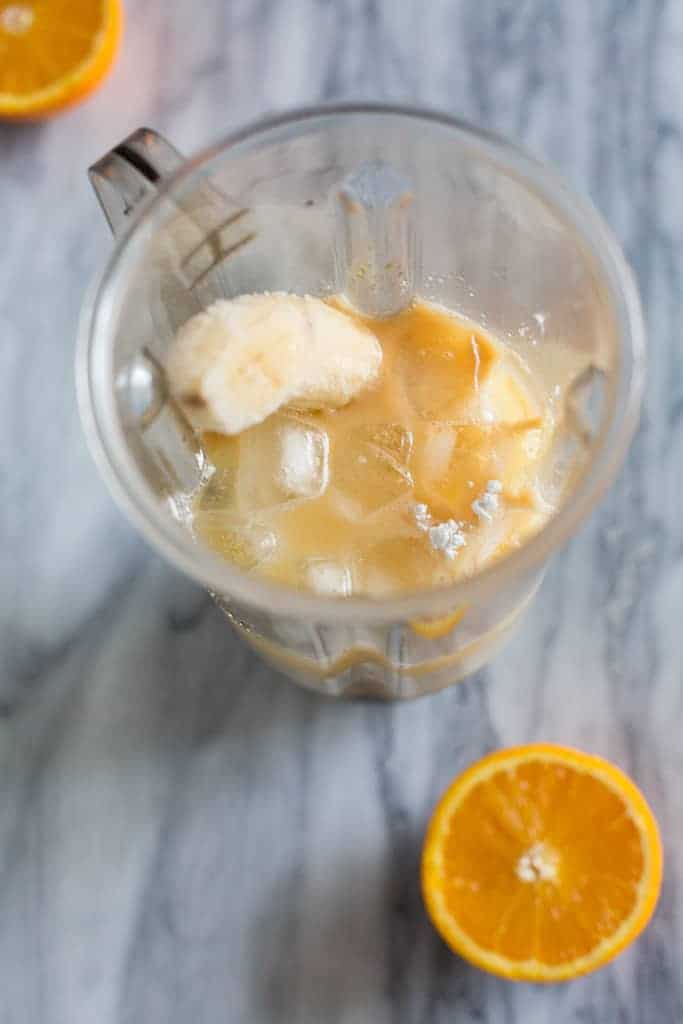 A blender with the ingredients for orange julius inside of it, including orange juice concentrate, ice, powdered sugar, vanilla, milk and a frozen banana.