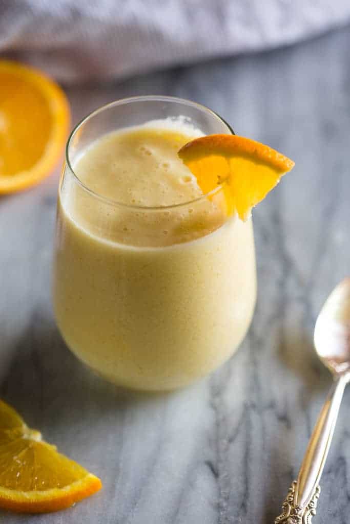 A clear glass of orange julius with a wedge of an orange on the rim and a spoon and sliced oranges in the background.
