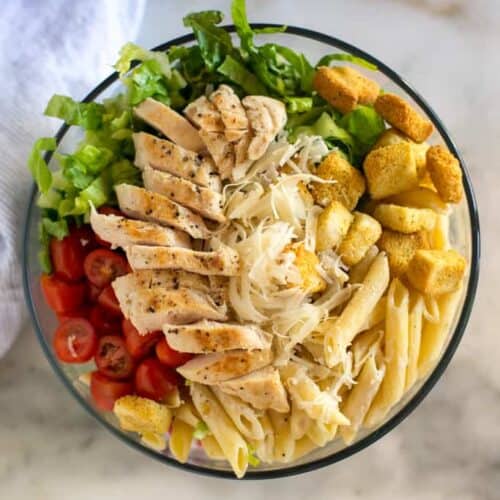 A bowl full of the ingredients to make chicken caesar pasta salad, including cooked penne pasta, chicken, tomatoes, croutons, lettuce and parmesan cheese.