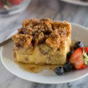 A slice of baked french toast casserole served on a white plate with syrup and berries.