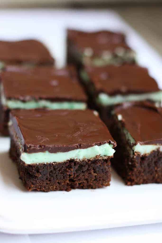 Chocolate brownies with a layer of green mint frosting, and melted chocolate on top, cut into squares and lined on a white tray for serving. 