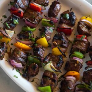 Steak Kebabs on wooden skewers with bell pepper and onion, served in a line on a white plate.