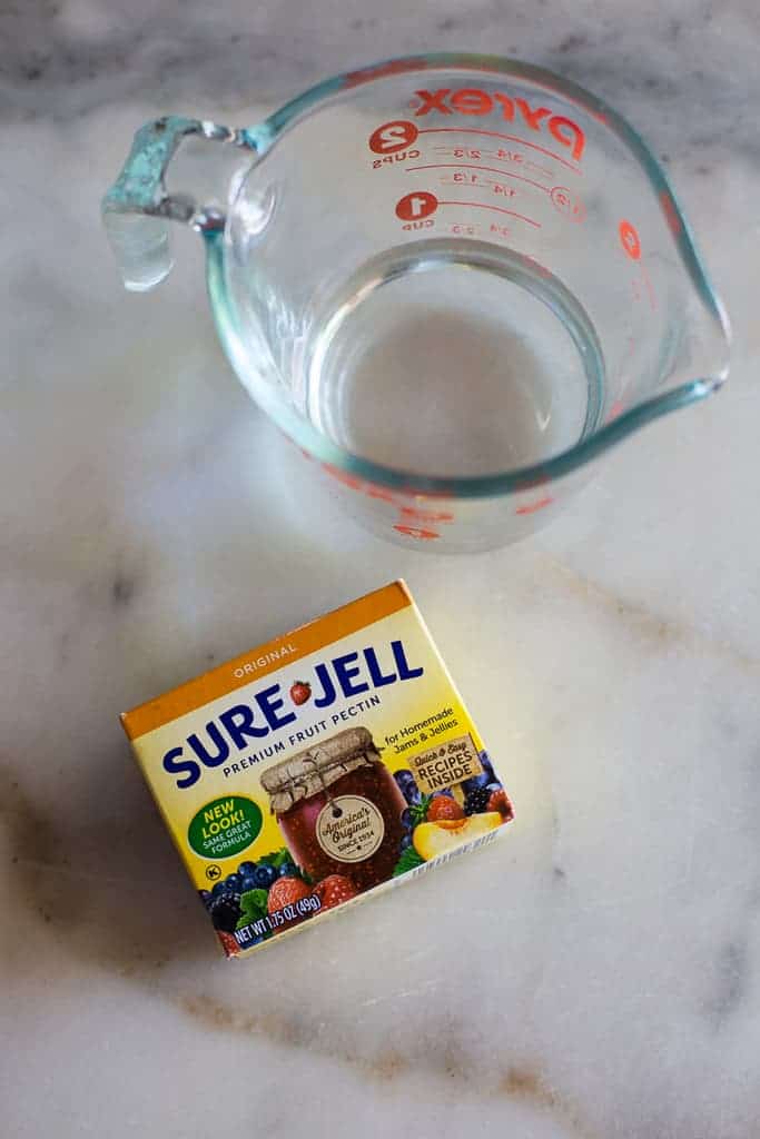A liquid measuring cup with water in it next to a package of Sure Jell fruit pectin.