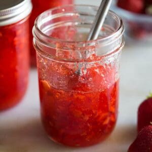 A mason jar half full of strawberry jam with a butter knife inside. Two additional jars of jam and fresh strawberries in the background.