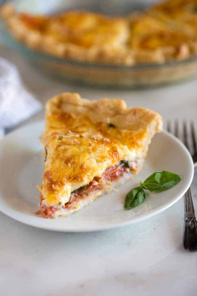 A slice of tomato pie on a white plate with a fork next to it and the rest of the tomato pie in the background.