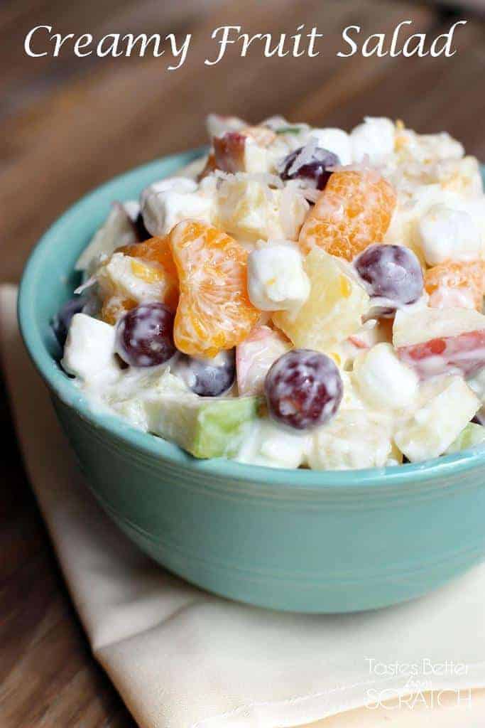 A blue bowl sitting on a napkin and filled with grapes, mandarin oranges, apples, marshmallows, and a Greek yogurt dressing.