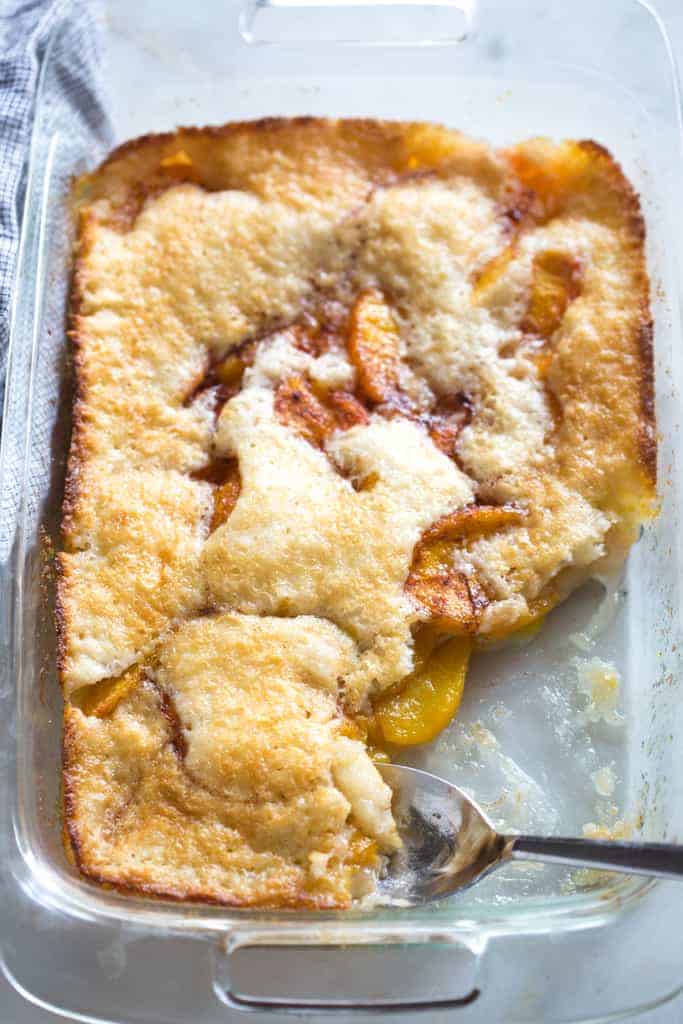 A 9x13 inch glass pan of peach cobbler with a spoon laying in the pan where a serving has been removed.